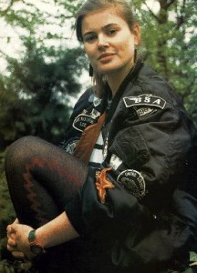 Ace: a young caucasian woman wearing a black bomber jacket covered in patches and badges. She is sitting in front of trees with her knee drawn up against her chest and is wearing black and red patterned tights. Image via Wikipedia, shared under Creative Commons licence