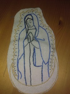 Photo showing an embroidered piece of white fabric on a wooden surface. The fabric is decorated with a traditionally styled image of the Virgin Mary in prayer, stitched in outlines of black, blue and gold. Photo by the author.