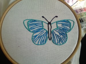 Photo of an embroidery hoop with a blue and green butterfly stitched onto the white fabric it supports. Photo by the author.