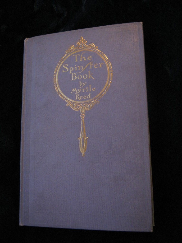 spinstercover An old clothback book. It is a lavender coloured hardback with a hand mirror inlaid in gold leaf on the front. The mirror has the text THE SPINSTER BOOK inside it. Image by the author.